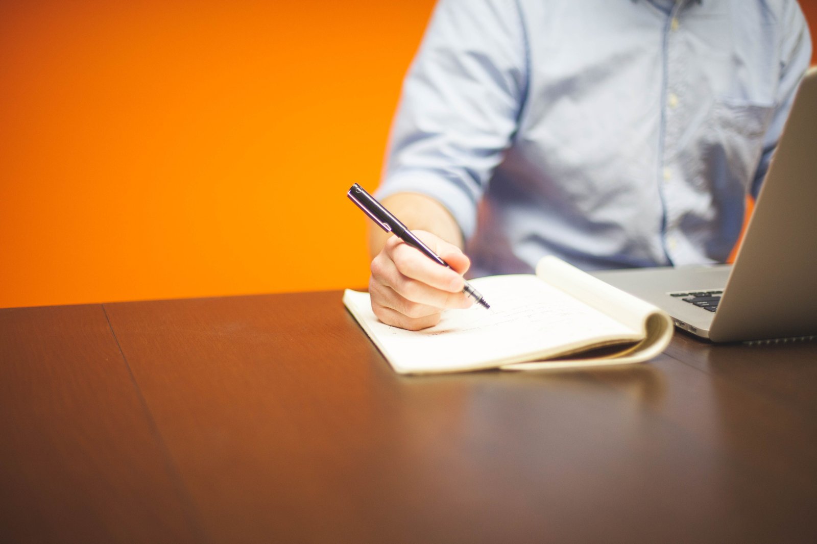 Persona escribiendo en un cuaderno junto a un portatil en un escritorio de oficina